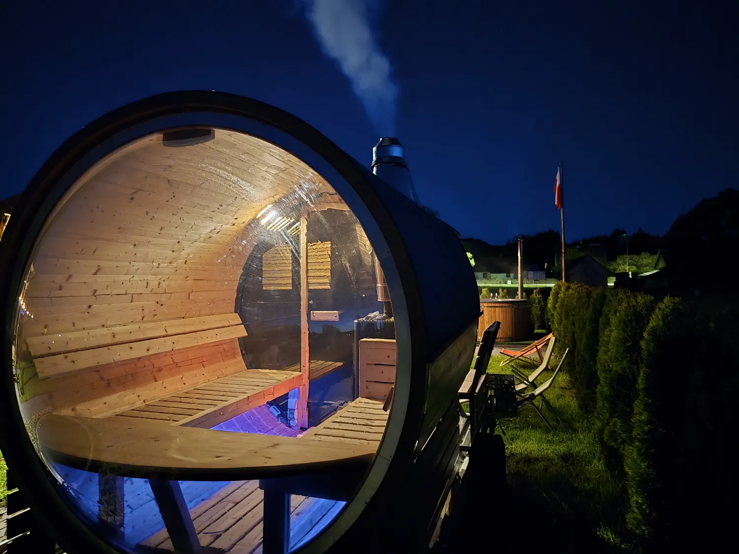 Rozgrzana sauna typu beczka wieczorem — rozświetlone wnętrze za panoramicznym oknem i dym unoszący się z komina